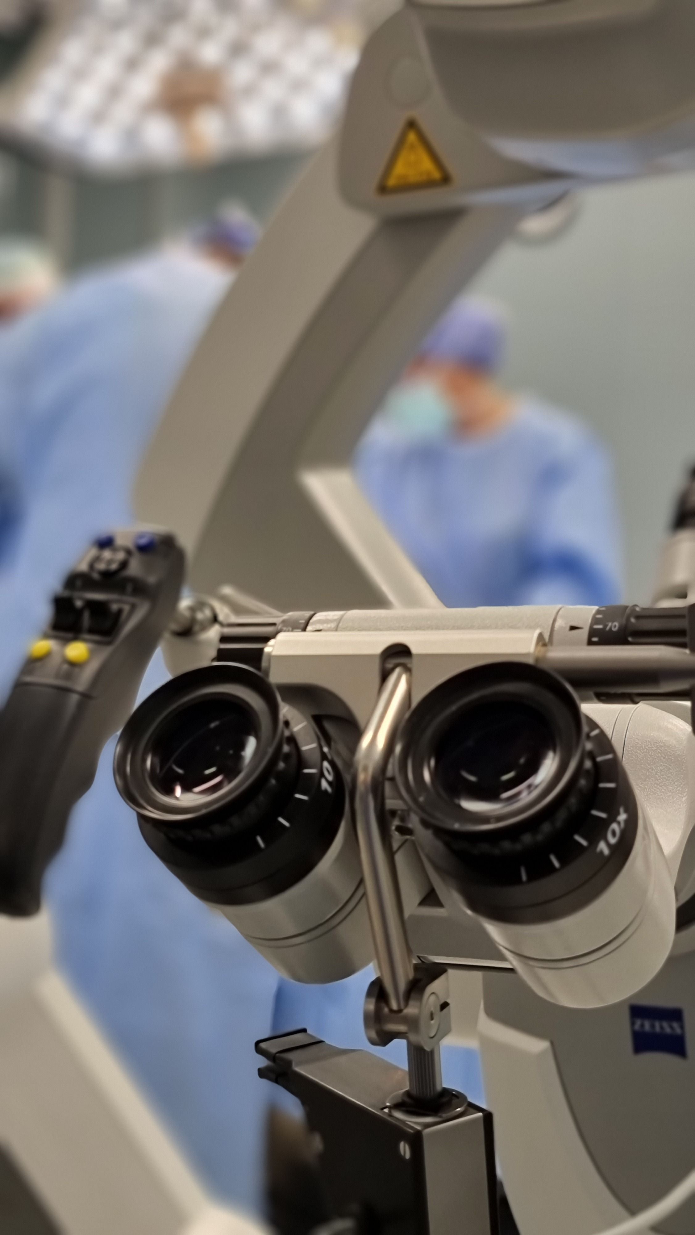 Prim-plan pe ocularele unui microscop chirurgical, cu echipa medicală în fundal.